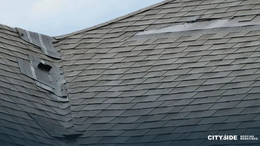 A close-up of a roof with a missing shingle and a shattered window