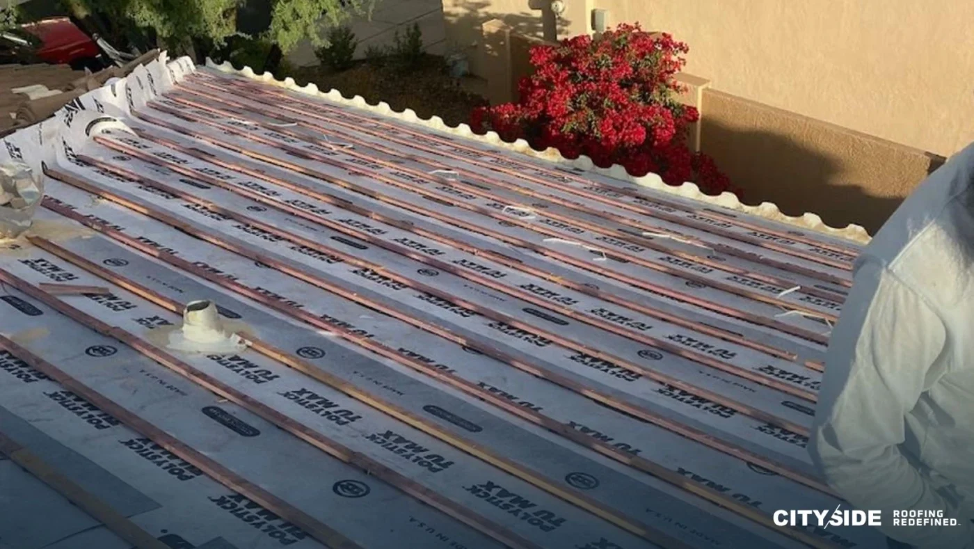 A man installs roofing materials on a house, standing on a ladder with tools in hand.