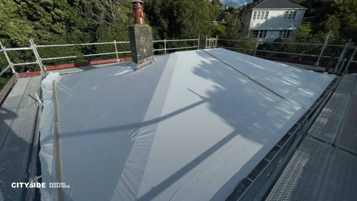 A flat roof covered in a white protective sheet surrounded by metal scaffolding. A chimney and nearby trees cast shadows, with a house in the background.
