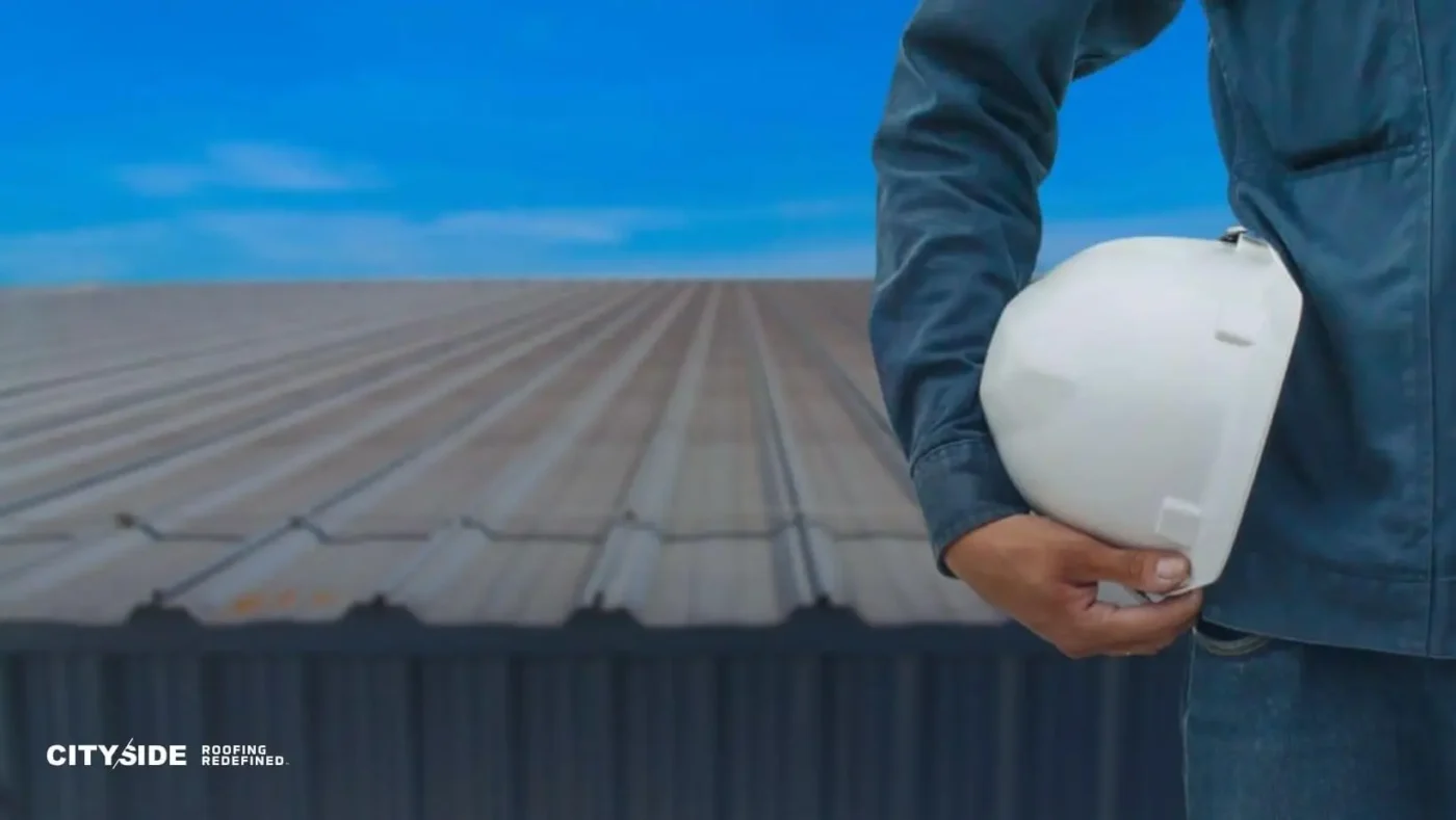 A person in a blue work jacket holds a white hard hat, standing on a corrugated metal roof under a clear blue sky, conveying readiness and professionalism.