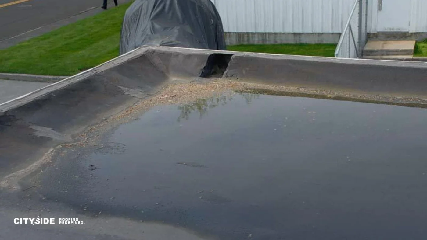 A flat rooftop with standing water pooling near a drainage area; a street, grass, and a covered item are visible in the background.