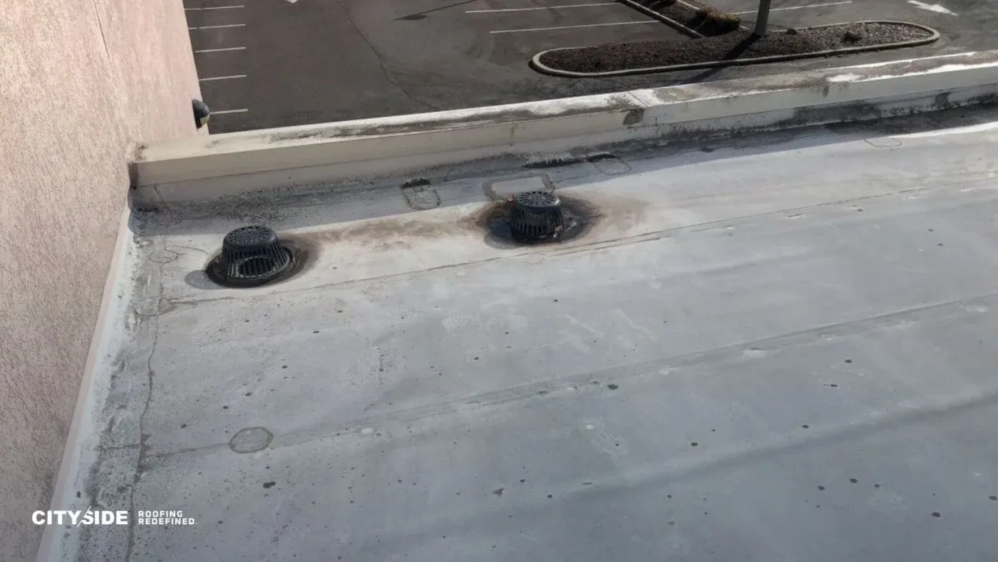 Flat rooftop with two circular vents, showing slight wear and dirt. Adjacent parking lot is visible, with a stark, utilitarian atmosphere.