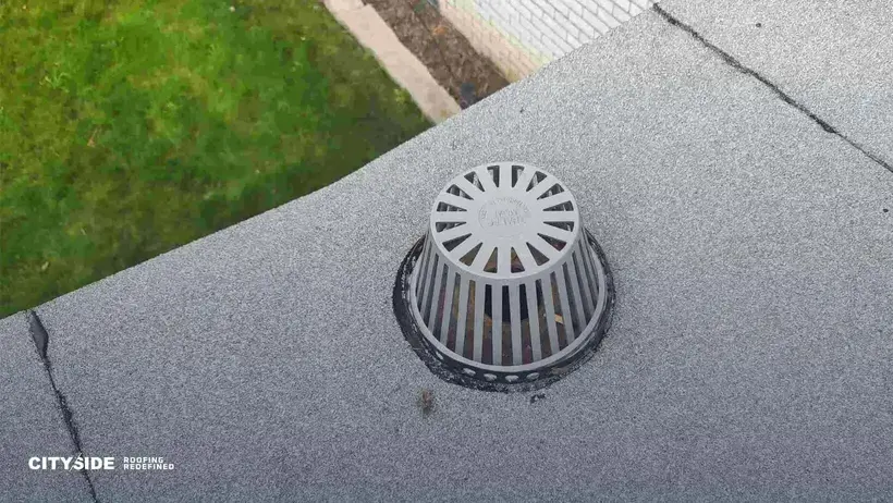 A roof vent on a flat surface, surrounded by grey roofing material and green grass in the background.