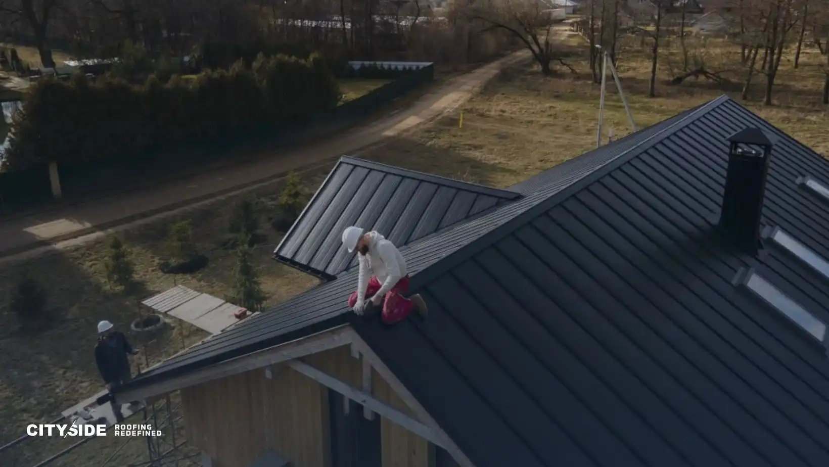 A person working on a dark, metal roof in a rural area, wearing safety gear. Another person stands below. The mood is diligent and focused.