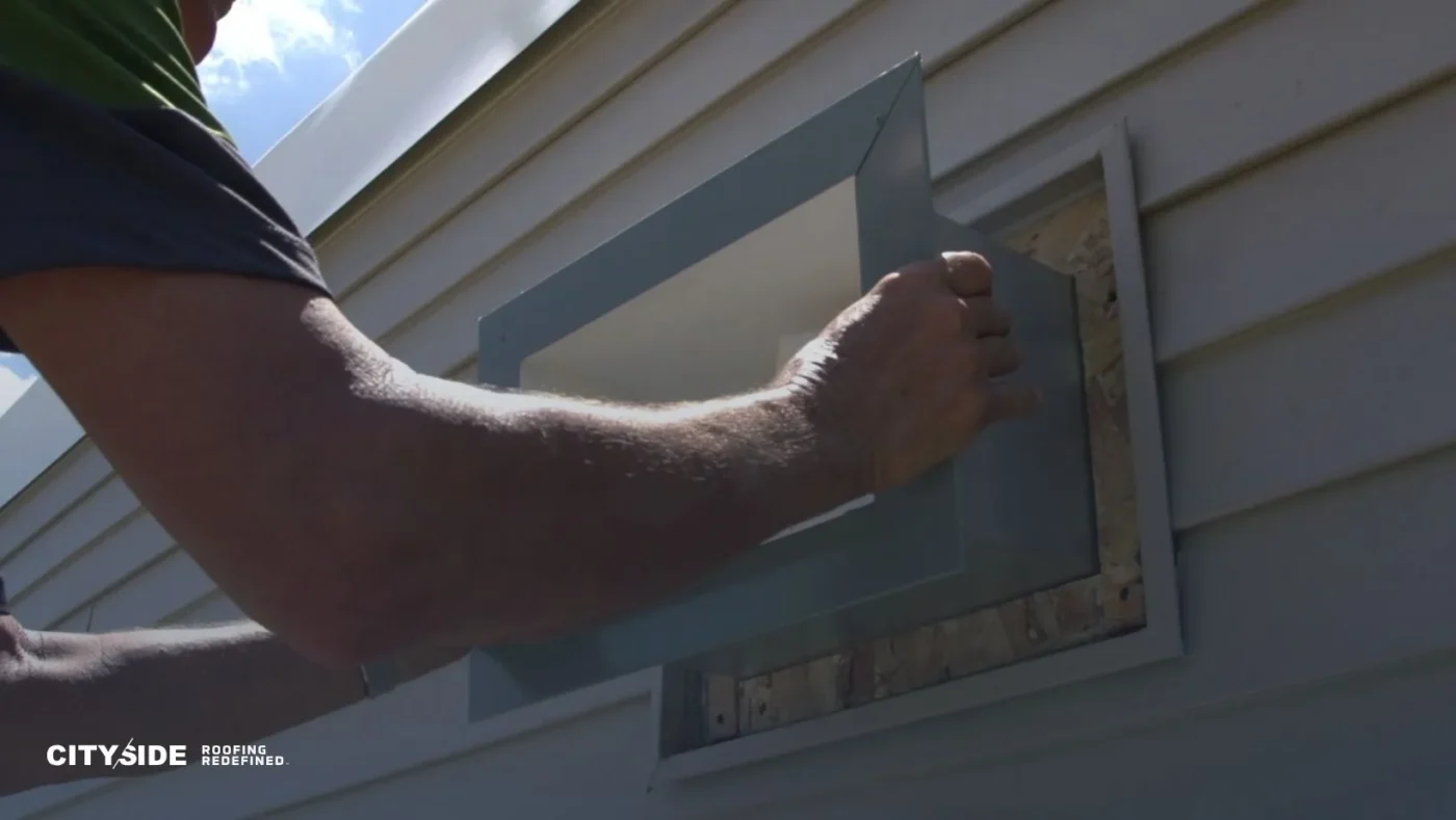 A person installs a gray vent on a house's beige siding under a blue sky. The image conveys a sense of professionalism and craftsmanship.