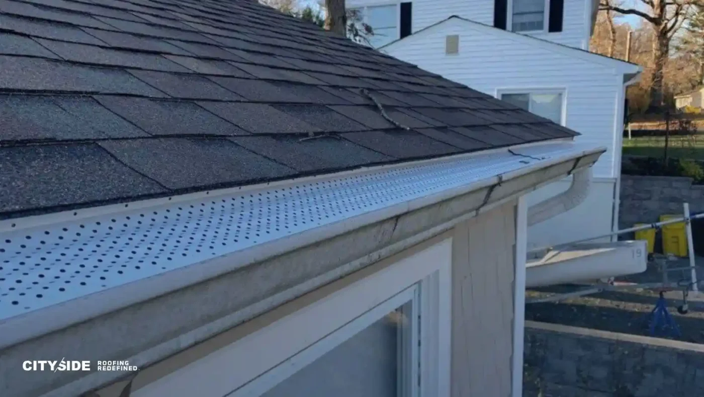 Close-up of a house roof with newly installed dark shingles and a perforated metal gutter guard. A white house is visible in the background.