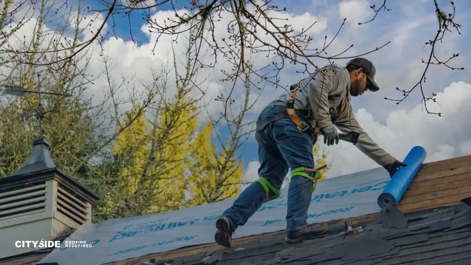 A worker in safety gear installs roofing material on a house under a blue sky. Leafless branches frame the scene, conveying a sense of focus and diligence.