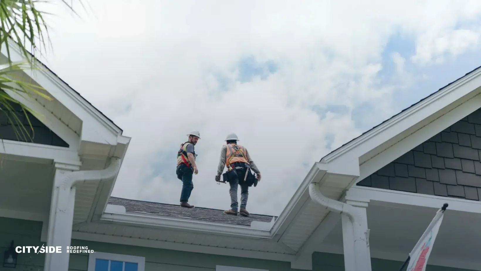 Two construction workers in safety gear stand on a house roof under a partly cloudy sky. They appear focused and are assessing the roofing work.