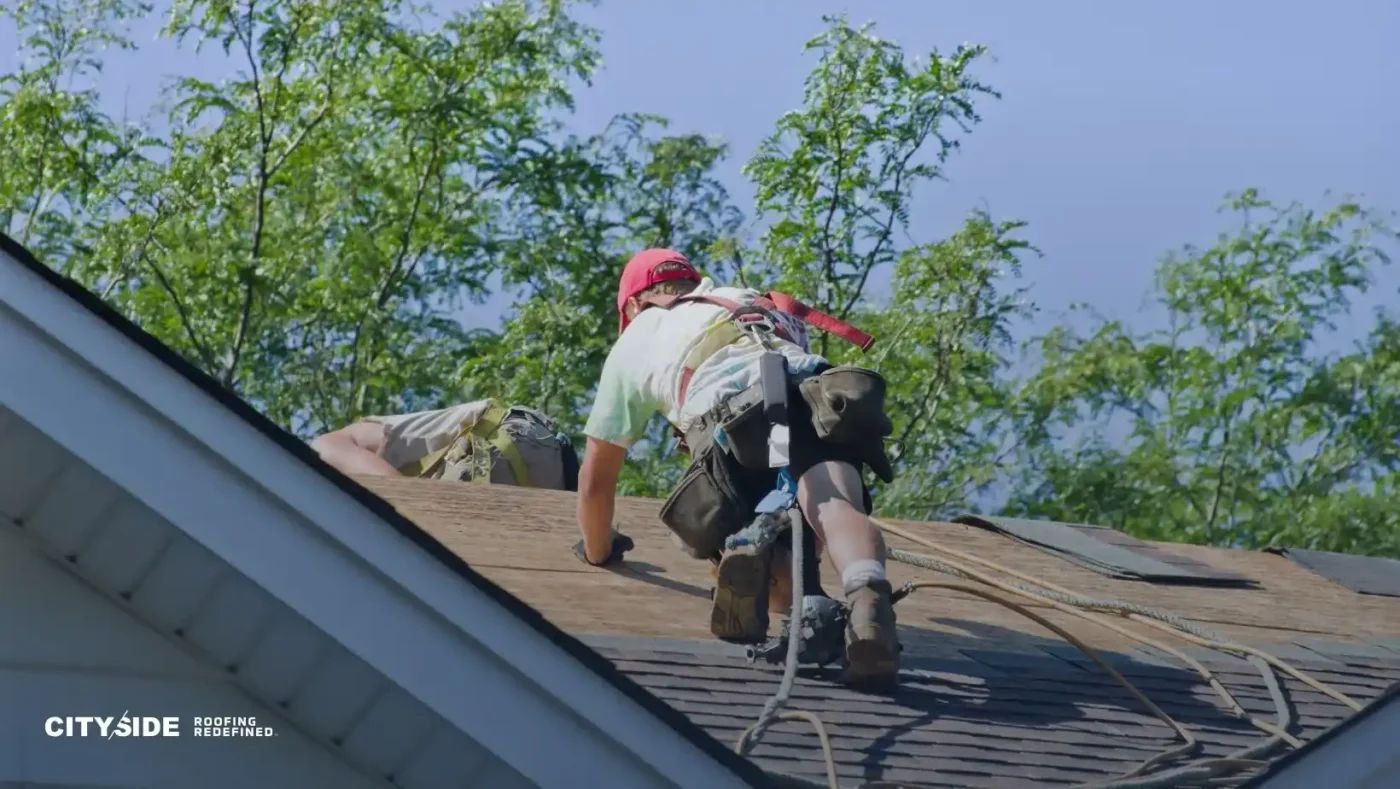 A worker in safety gear and a red cap repairs a roof on a sunny day. Trees and blue sky form the background. The scene conveys focus and diligence.