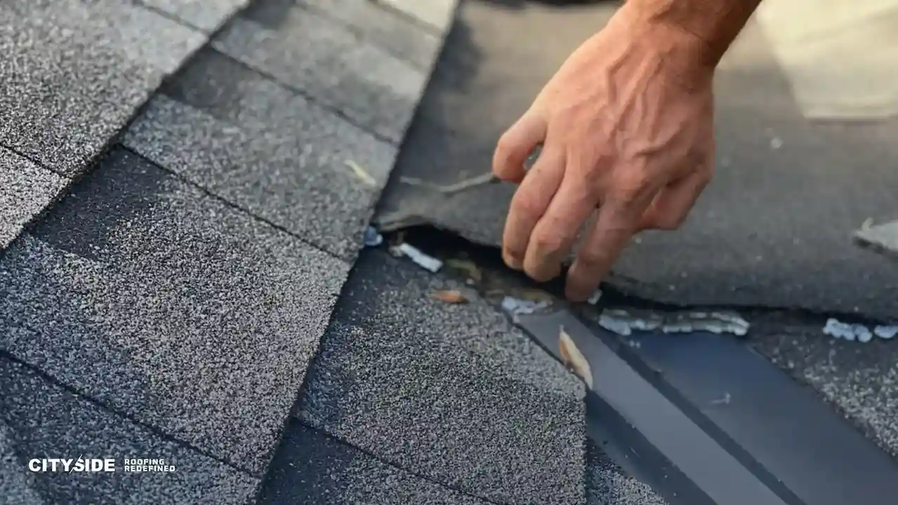 A hand inspects damaged asphalt shingles on a roof, revealing several bent and broken pieces. The scene conveys concern and the need for repairs.