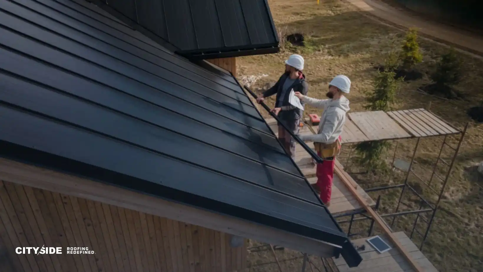 Two workers in hard hats inspect a modern metal roof on a wooden house. Scaffolding surrounds the building, suggesting ongoing construction.