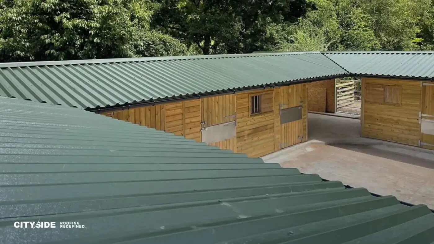 Green metal-roofed stables form a U-shape against a backdrop of lush trees. The scene conveys a serene, rustic atmosphere.