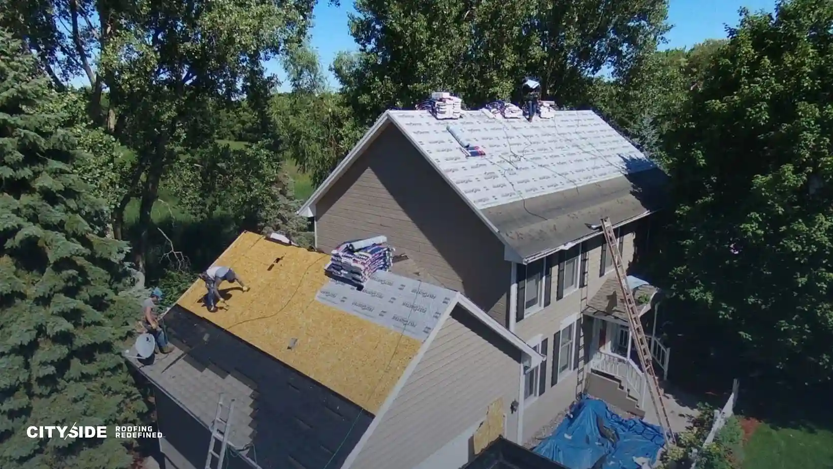 Aerial view of roofers working on a house with trees surrounding it. One section is covered in tar paper, while another side is being prepared.