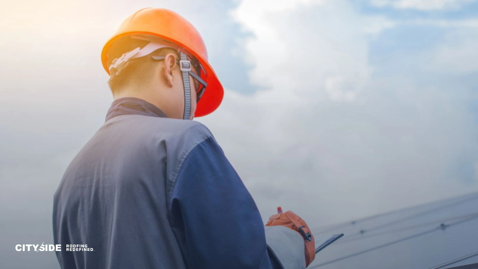 A worker in an orange hard hat and gray jacket is examining a roof with a tablet, under a cloudy sky. Text reads "Cityside Roofing Redefined."