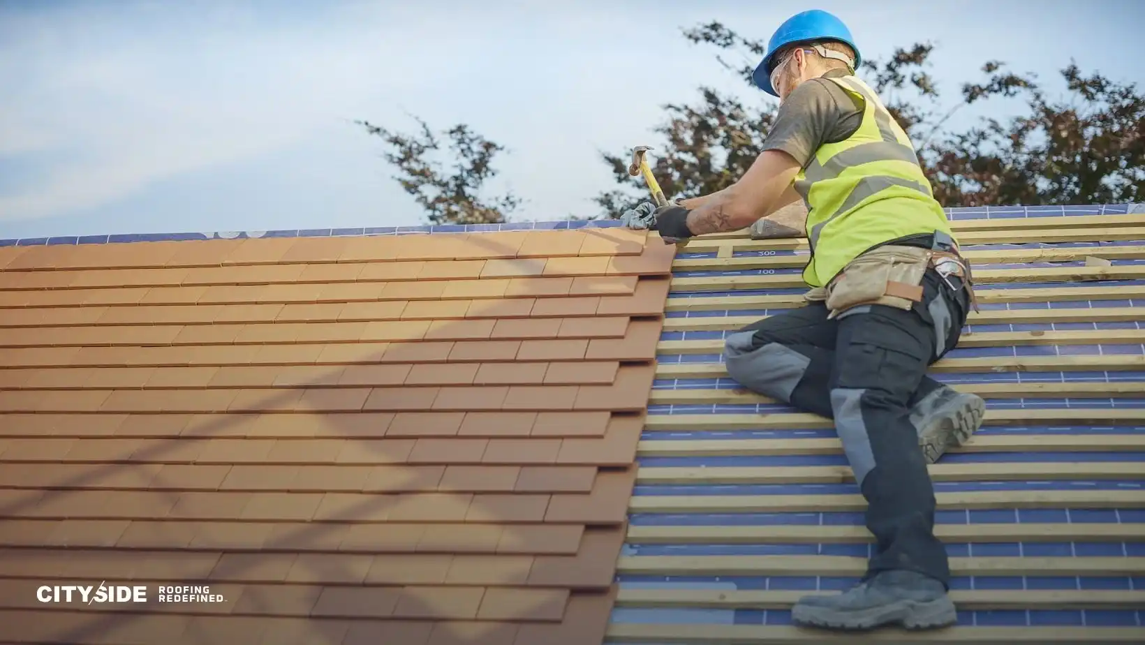 A construction worker in a blue helmet and yellow vest installs shingles on a roof. The worker holds a hammer, with trees in the background. The atmosphere is focused and professional.