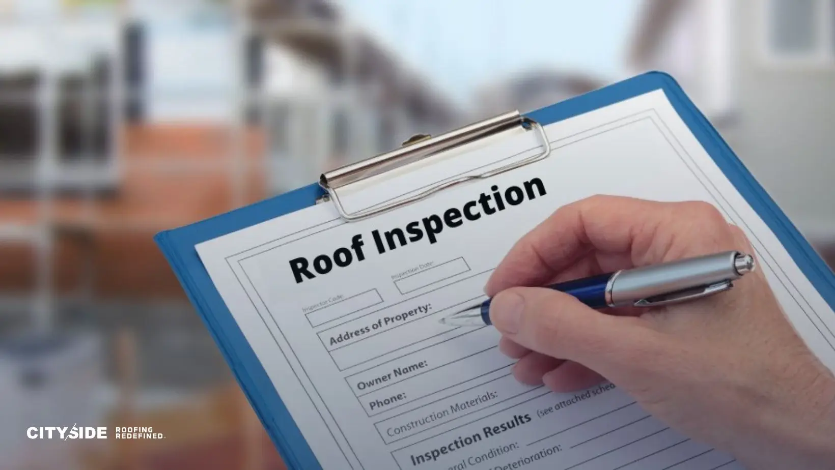 A person holds a pen poised to write on a roof inspection form attached to a clipboard. The background shows a blurred image of houses, suggesting a residential area.