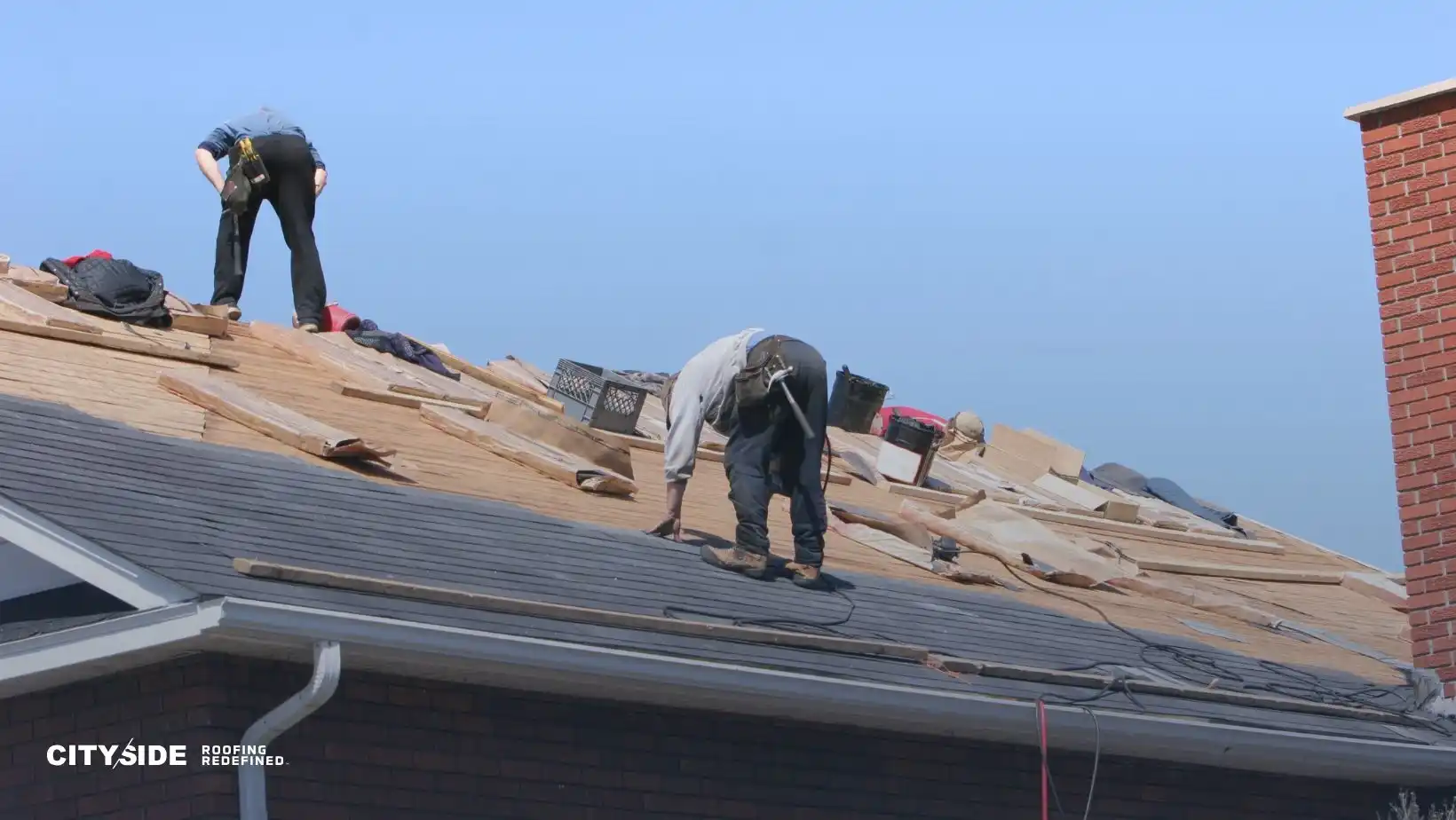 Two workers are reroofing a house, surrounded by tools and wooden planks, under a clear blue sky. The scene conveys industriousness and teamwork.