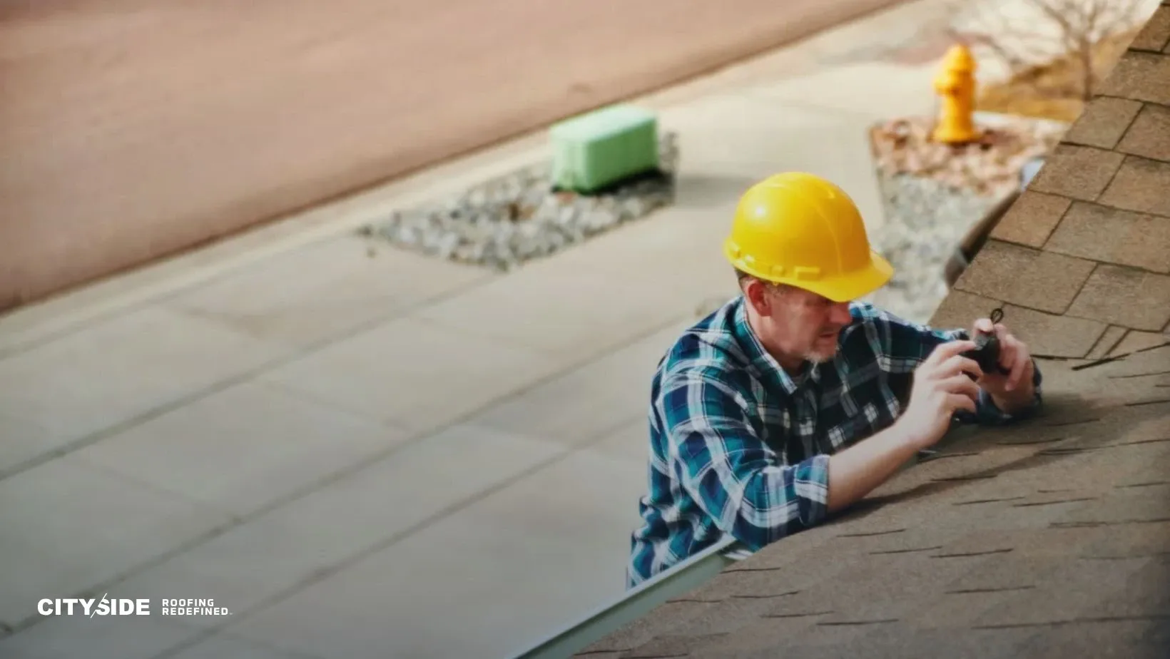 Man in a yellow hard hat and plaid shirt inspecting a roof. He holds a tool and appears focused. A sidewalk and fire hydrant are visible in the background.
