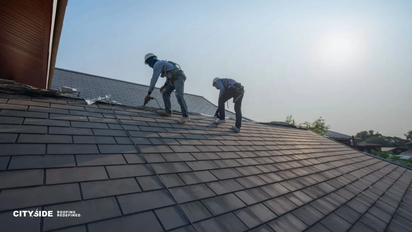 Two workers in safety gear repair a sloped roof under a clear sky. The scene conveys teamwork and precision. Sunlight casts long shadows.