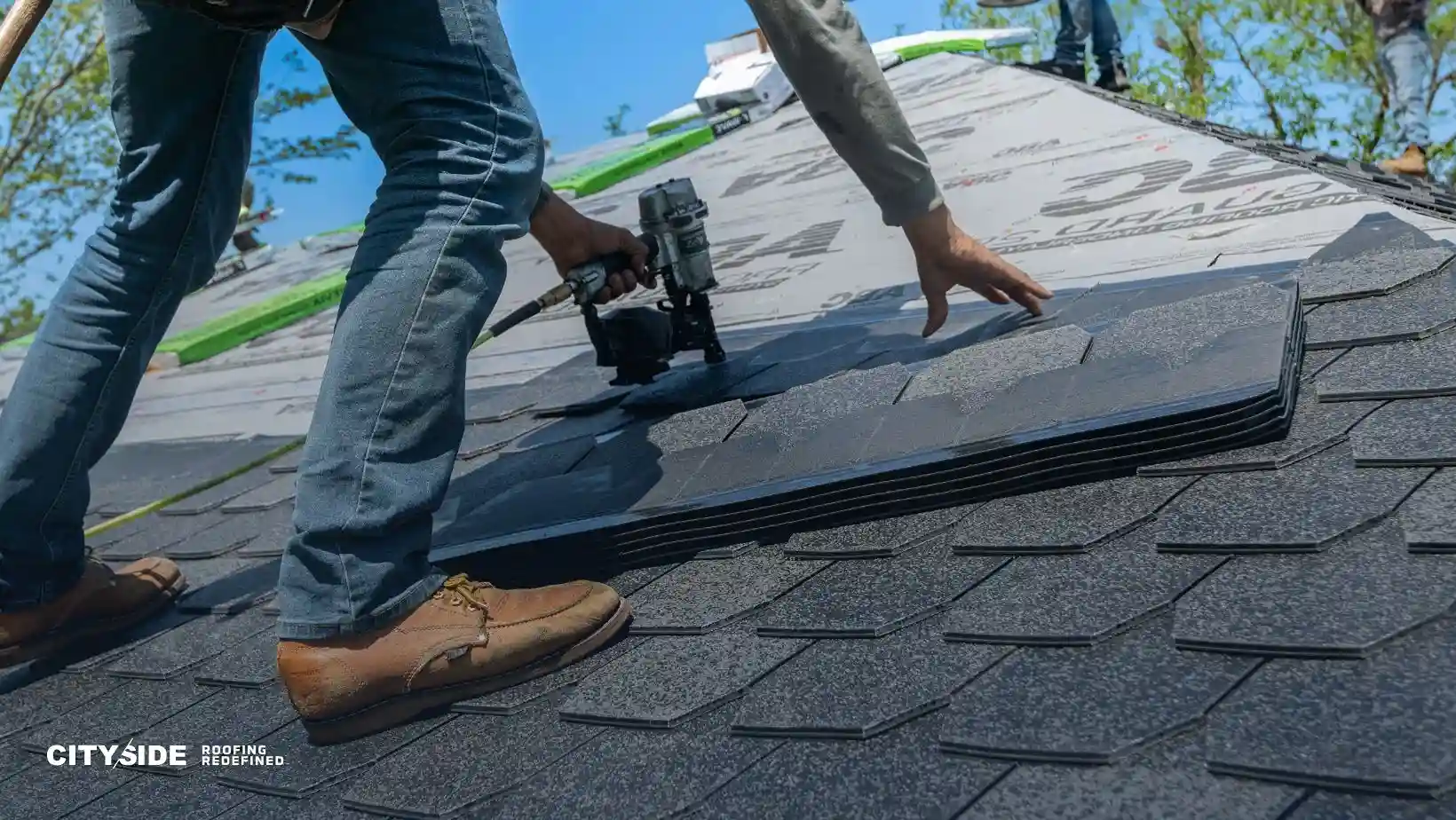 A worker in jeans and boots installs dark shingles on a sloped roof using a nail gun. The scene conveys a sense of precision and craftsmanship.