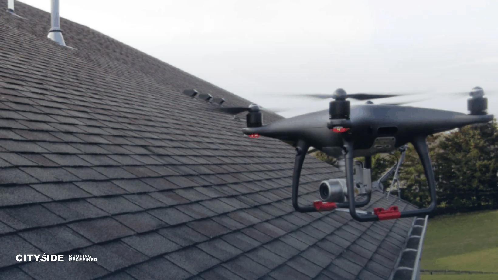 A black drone hovers close to a sloped, shingled roof, likely inspecting its condition. The background reveals a blurred line of trees and an overcast sky.