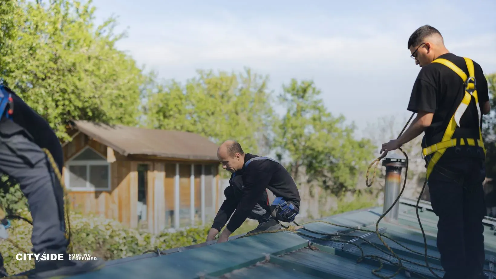 Three workers in safety gear are installing a metal roof on a house. The setting is outdoors with trees and a cabin in the background. The mood is focused.