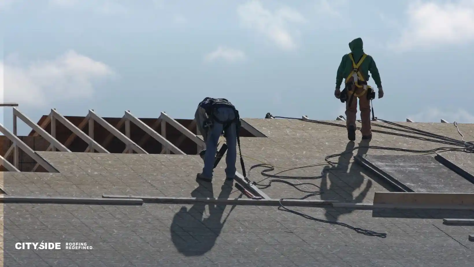 Two workers in safety gear are on a roof under construction against a cloudy sky. One is bending over, working, while the other stands, observing.