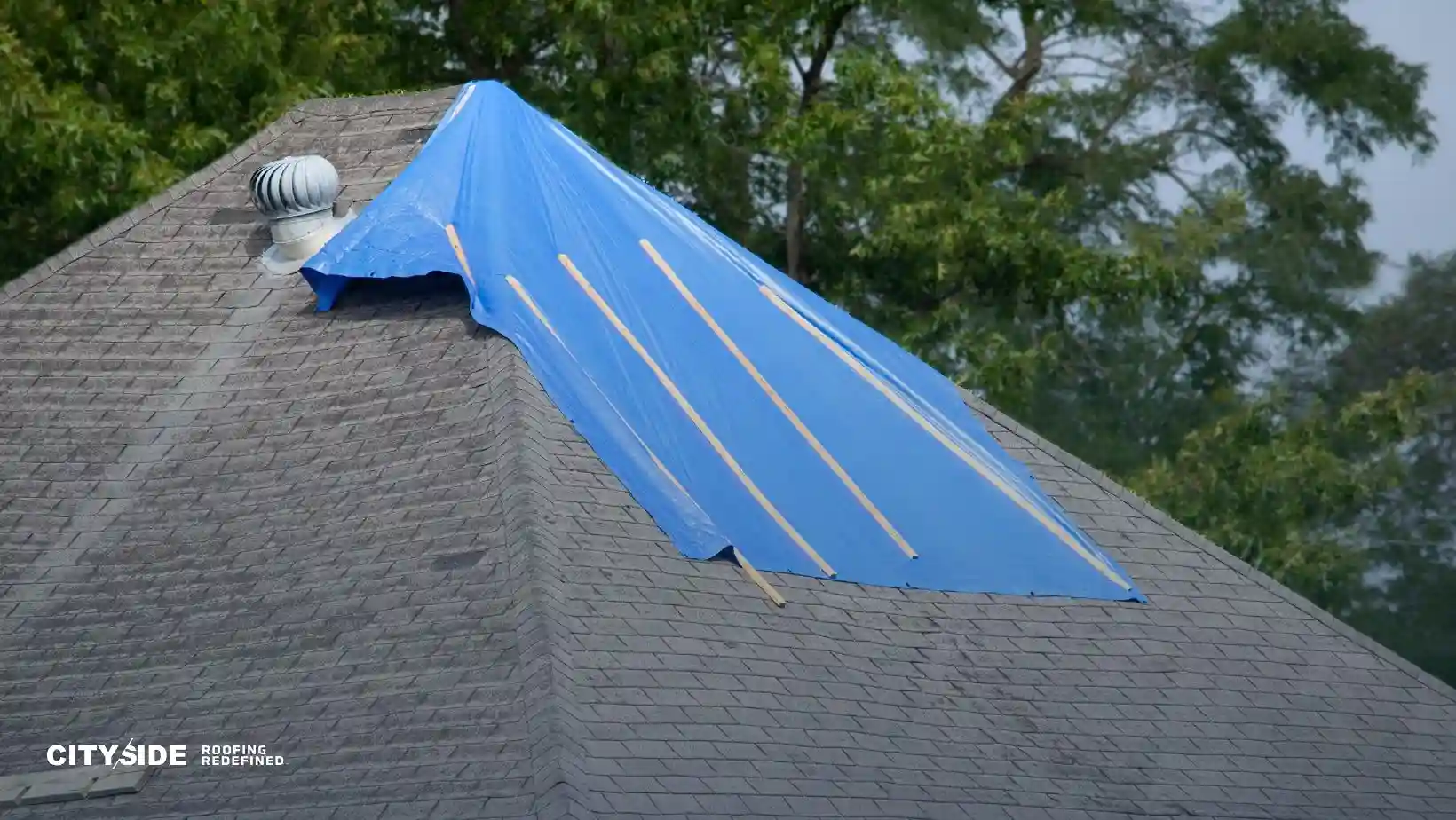A roof with a blue tarp covering a damaged section, secured with wooden planks. A white vent is visible. Trees in the background; overcast atmosphere.