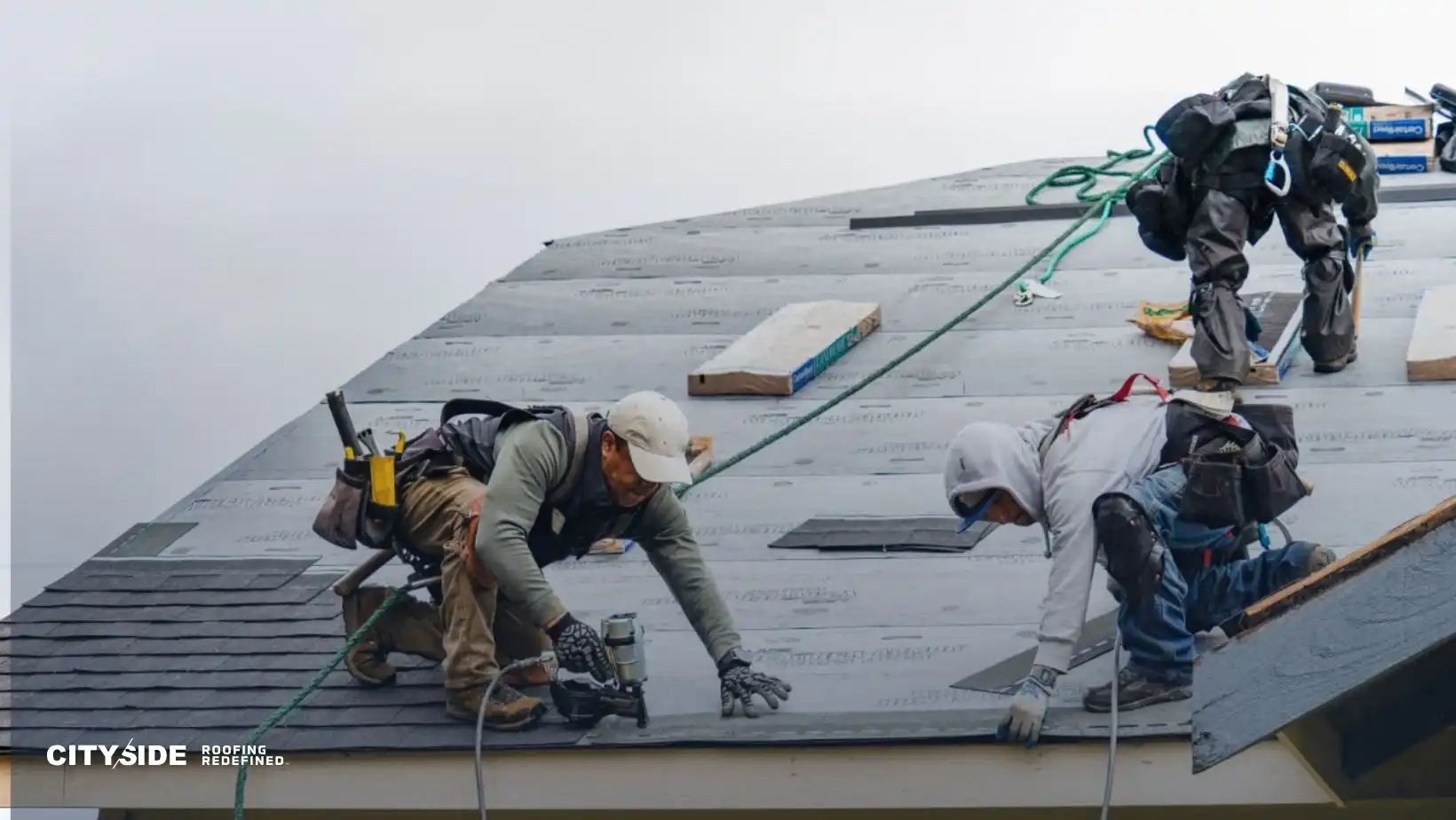 Two roofers in protective gear work on shingling a house roof. They are focused and using tools, conveying teamwork and precision. Overcast sky.