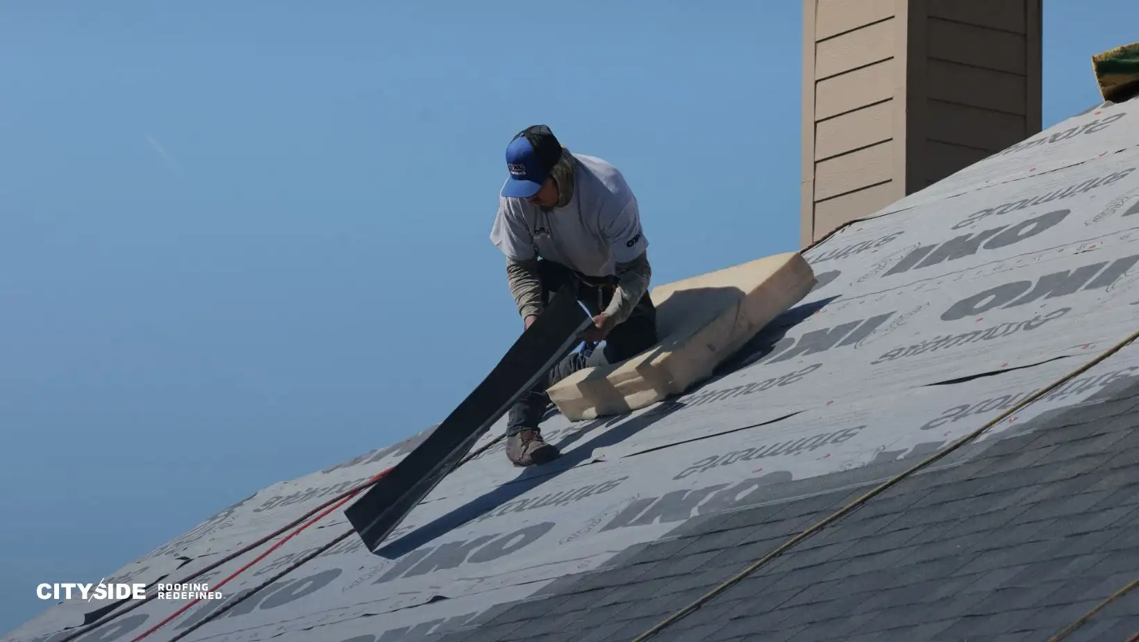 Man wearing a blue cap and gray shirt working on a steep roof under a clear blue sky, handling roofing materials. "Cityside Roofing" logo in the corner.