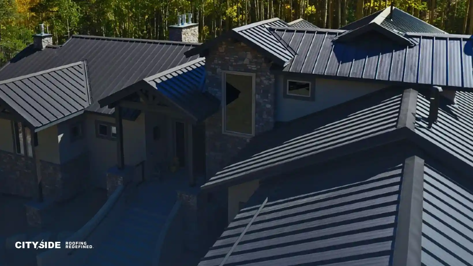 Aerial view of a modern house with sleek, dark metal roofing. Surrounded by trees, it conveys a serene and contemporary architectural style.