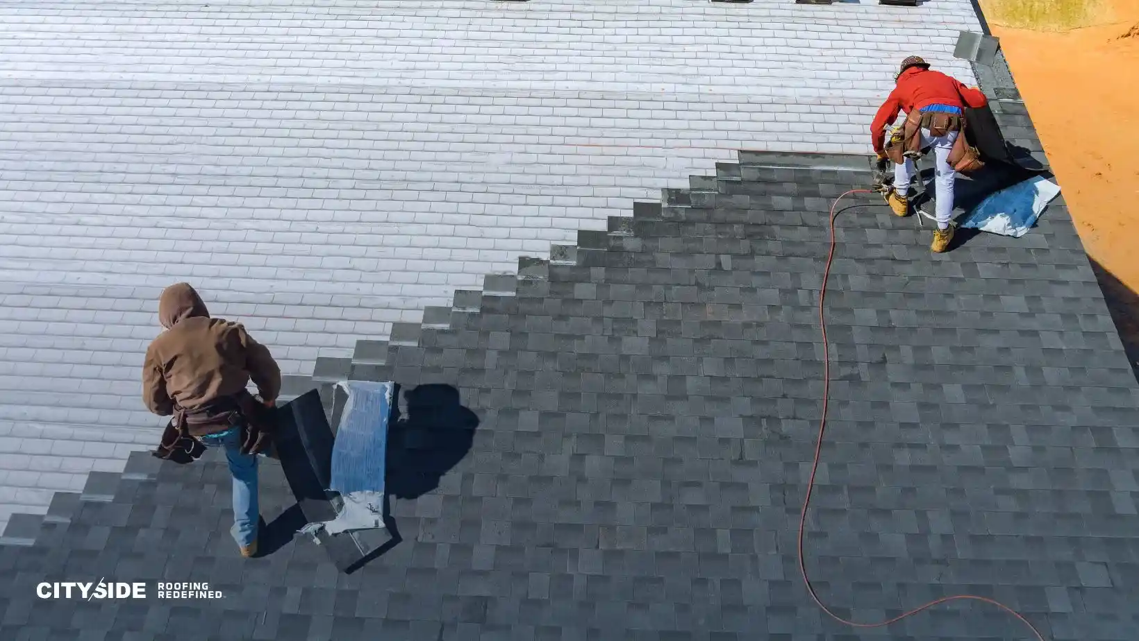 Two construction workers in hoodies install dark gray shingles on a roof, one crouched with a tool, conveying teamwork and focus under a clear sky.