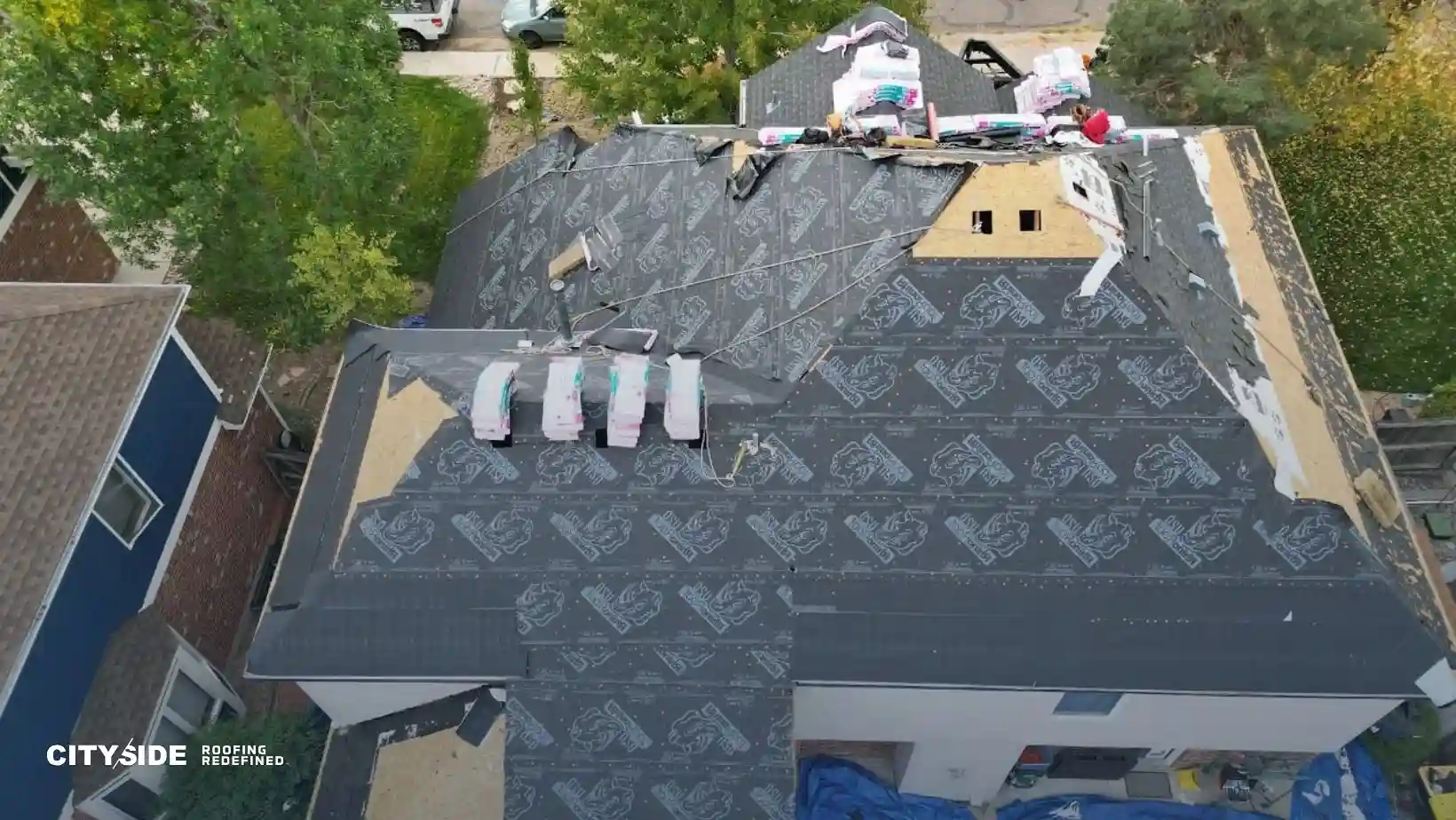 Aerial view of a house roof under construction, covered in protective underlayment. Roofing materials are stacked, surrounded by green trees.