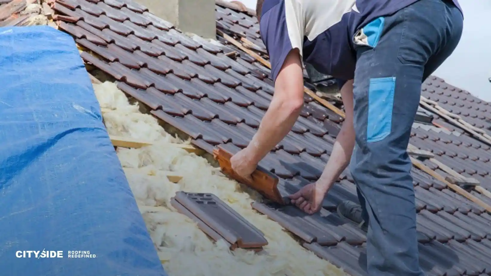 A worker in blue pants repairs a tiled roof, securing tiles and adjusting insulation. A blue tarp covers part of the roof. The scene conveys focus and craftsmanship.