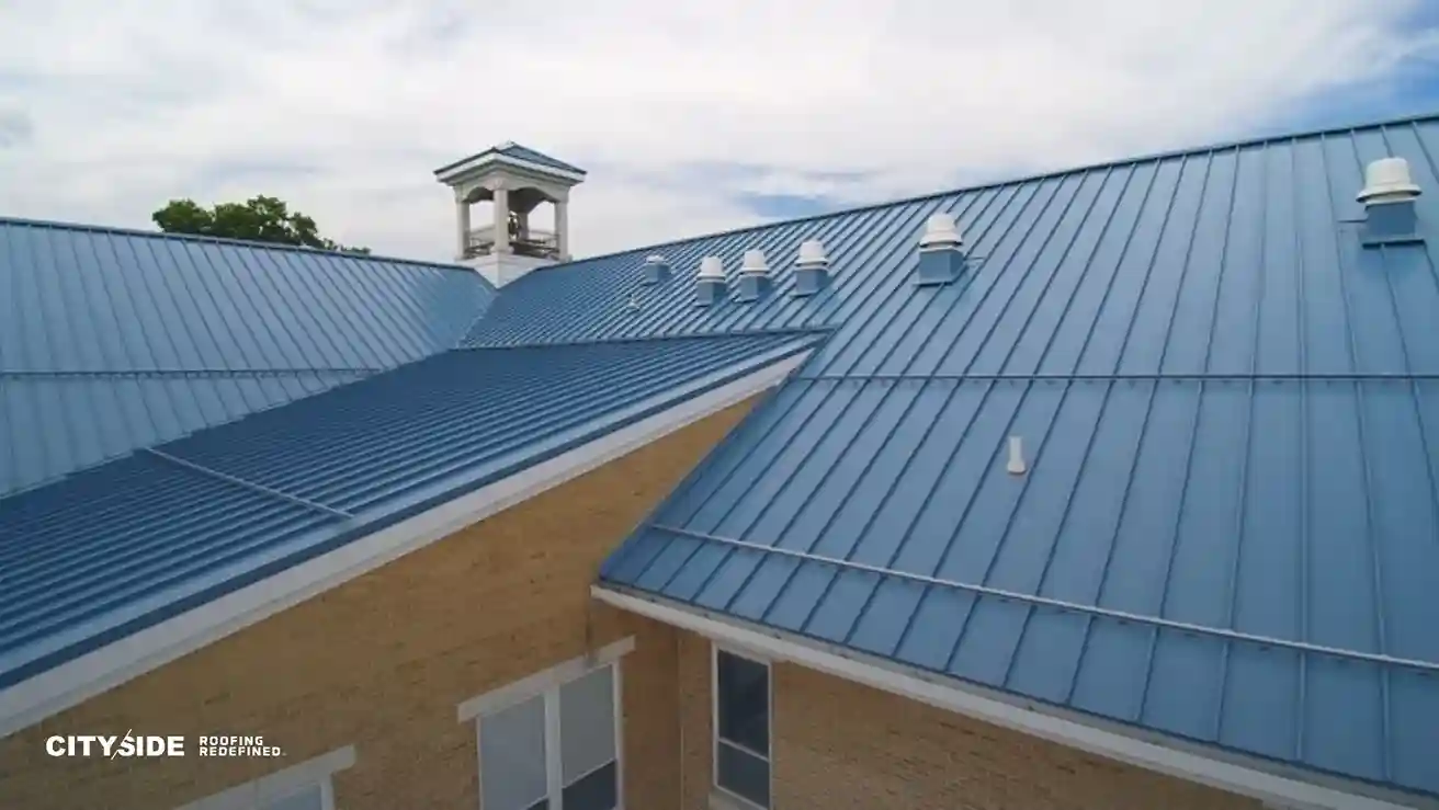 Blue metal roof of a beige brick building under a cloudy sky. The roof features neat rows of vent stacks. Logo reads "Cityside Roofing Redefined."