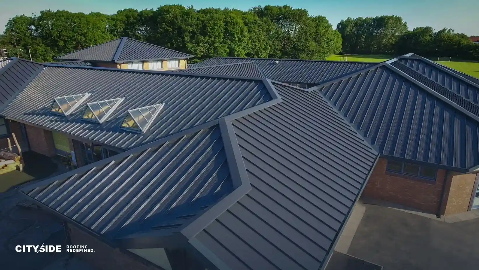 Aerial view of a modern building with a sleek, dark metal roof featuring angular designs and skylights. Surrounded by lush green trees under a clear sky.